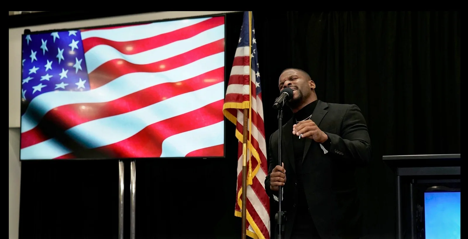 A singer performs the national anthem from the Business Expo stage in front of a large screen showing the American flag, with a flag standing beside him.