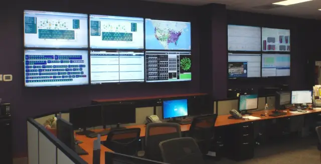 Control room with multiple monitors displaying data charts, maps, and graphs, featuring workstations with computers and office chairs.
