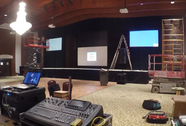 A stage setup with lighting and audio equipment in an auditorium, featuring a scissor lift, a ladder, and screens displaying test patterns.