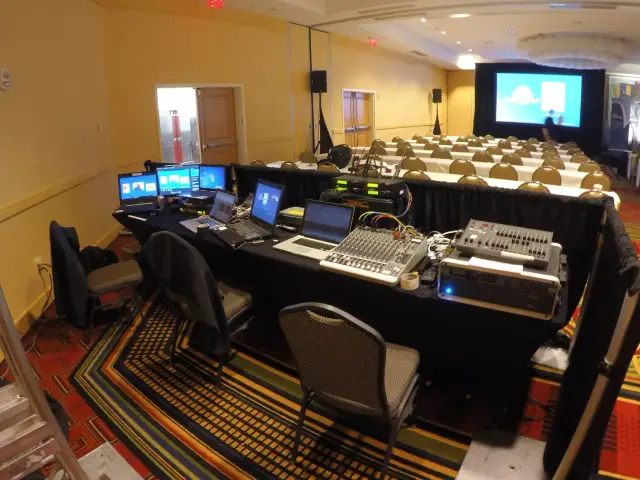 Audio-visual setup in a conference room with laptops, monitors, and audio equipment, facing rows of chairs and a presentation screen.
