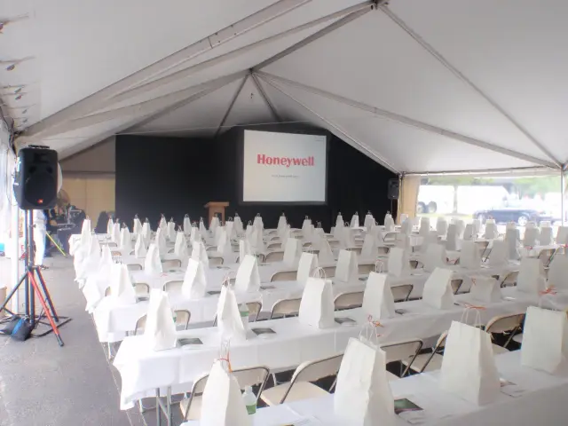 Conference setup under a white tent with rows of chairs and tables, each table holding a white gift bag and documents. A podium and a screen displaying the Honeywell logo are at the front.