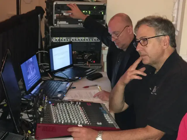 Two men operate audio and visual equipment, including laptops and a sound mixer, in a tech setup.