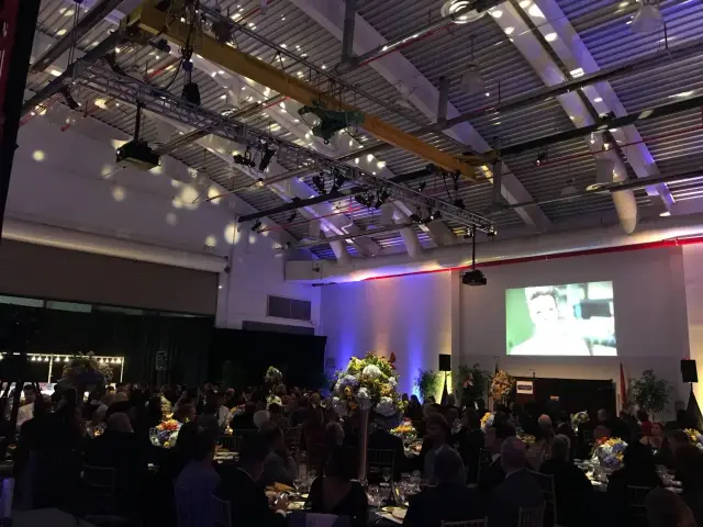 Indoor event with attendees seated at tables, floral centerpieces, large screen displaying a presentation, and overhead lighting fixtures.