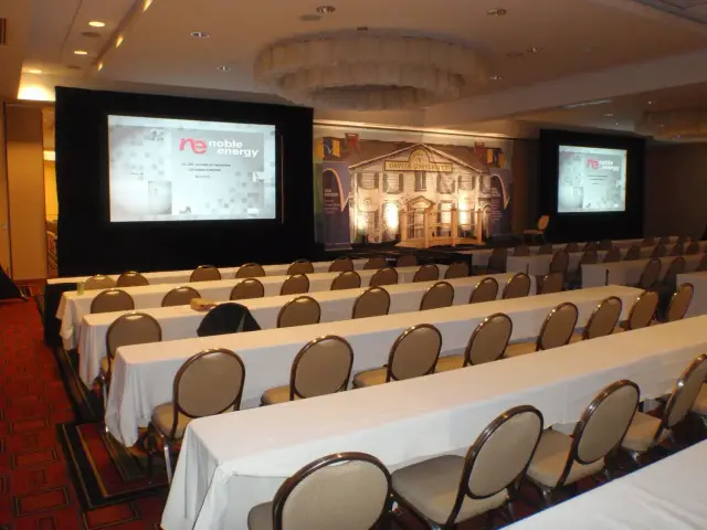 Conference room with rows of tables and chairs facing two projection screens displaying "Noble Energy" presentation, with a backdrop labeled "Davita.