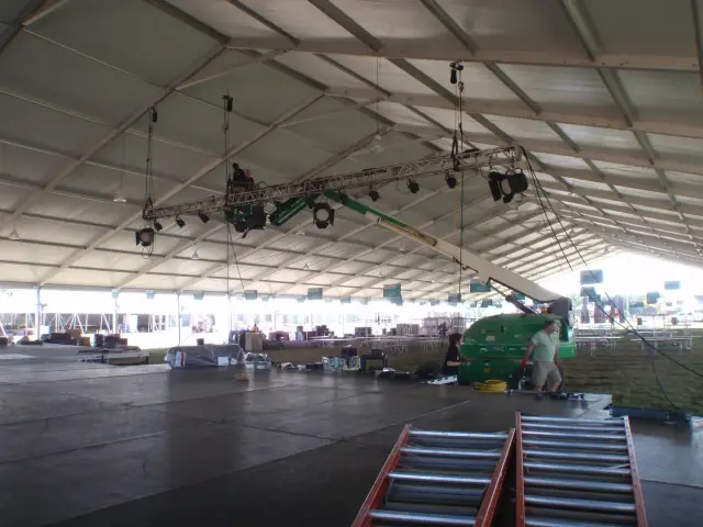 Interior view of a large event tent with construction equipment and workers setting up lighting rigging from a lift.