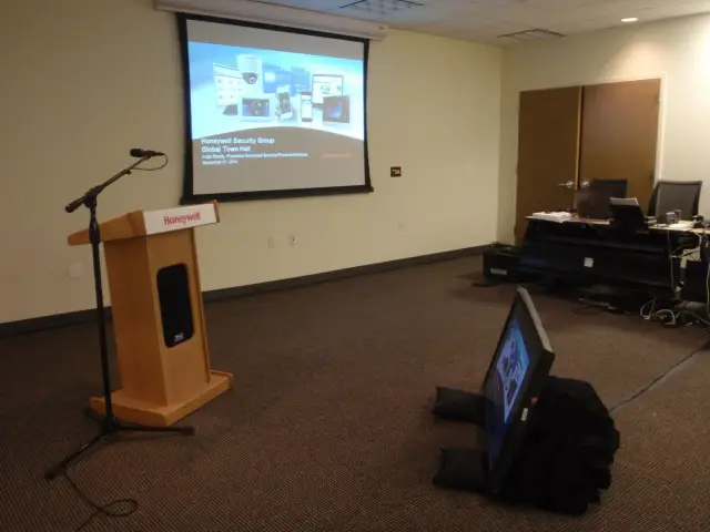 Conference room with a podium and a projection screen displaying "Honeywell Security Group Global Town Hall." A microphone is on the podium, and a monitor lies on the ground.
