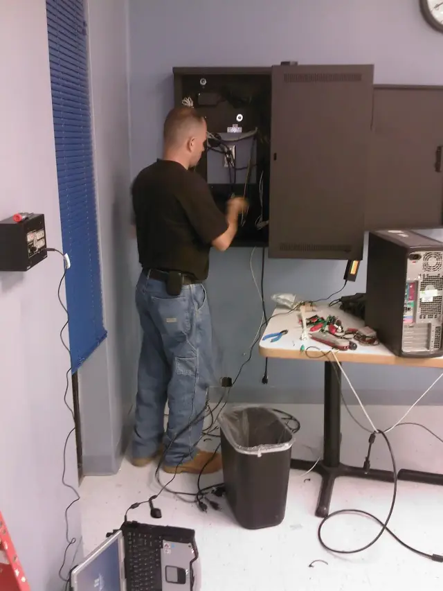 Person working on electronic equipment in a room, with cables and tools on a table nearby.