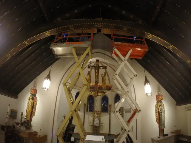 Church interior with dark wooden vaulted ceiling, ornate decorations, crucifix above altar, religious statues, and two scissor lifts with workers near the ceiling.