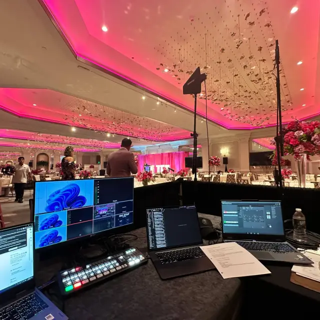 Production table at a Garden City Hotel gala: a video switcher, multiview monitor and laptops facing the ballroom, with wireless microphone antennas on stands and the stage lit pink in the distance.