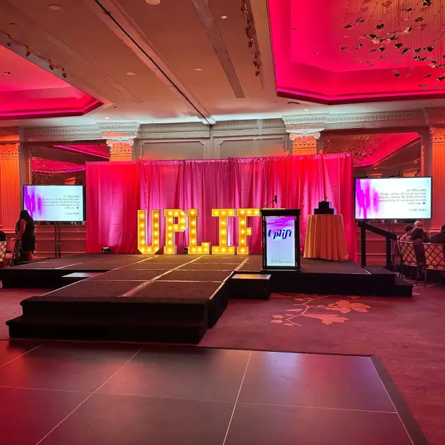 Gala stage in a Garden City Hotel ballroom: lit marquee letters spelling UPLIFT in front of a pink-lit drape, a branded podium, and a display screen on each side of the stage.