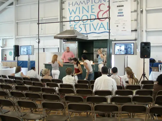 Indoor cooking demonstration at Hampton Wine & Food Festival with audience seated and a presenter on stage.
