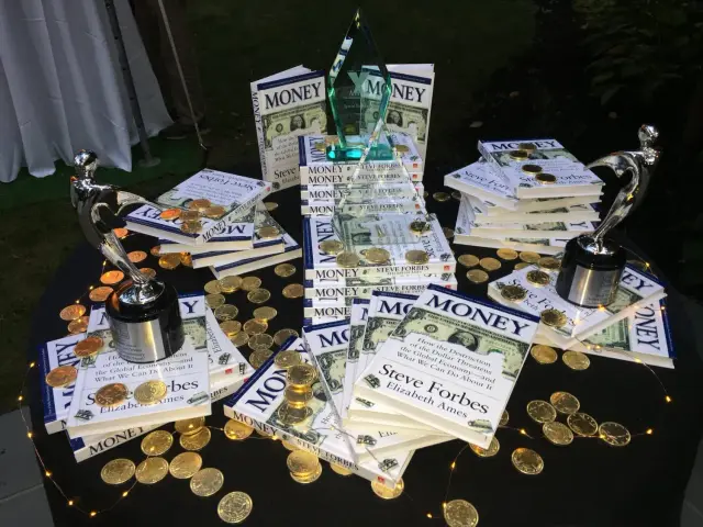 A table displaying stacks of books titled "Money" by Steve Forbes and Elizabeth Ames, decorated with gold coins and fairy lights. The table includes three silver trophies and a glass award.