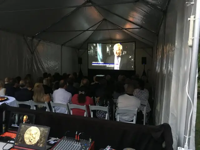A group of people seated inside a tent watching a video projected on a screen. The setting appears to be an event or presentation with audio-visual equipment and a laptop present.