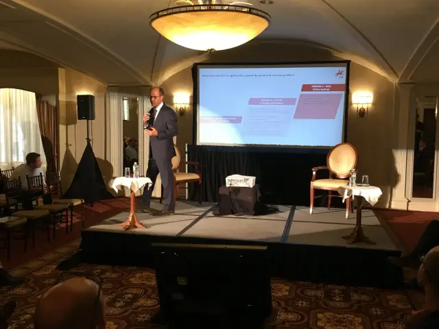 A speaker in a suit giving a presentation on stage with a projector screen behind him in a conference room.