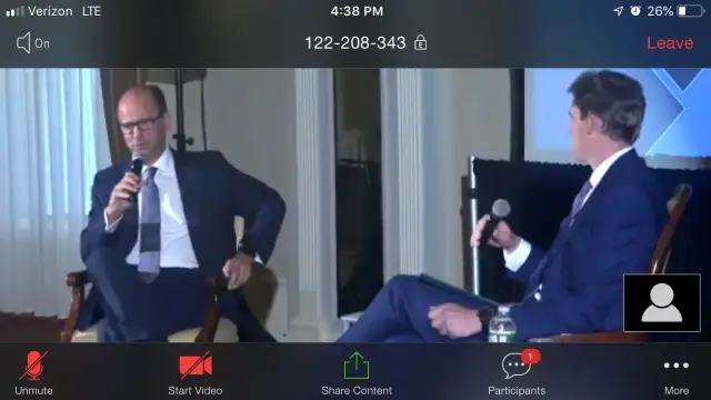 Two people in suits holding microphones during a video call conference, seated across from each other in a formal setting.