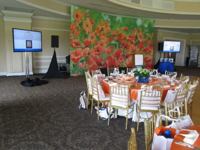 Banquet room setup with round table, orange tablecloth, and elegant chairs, large floral backdrop, and presentation screens.
