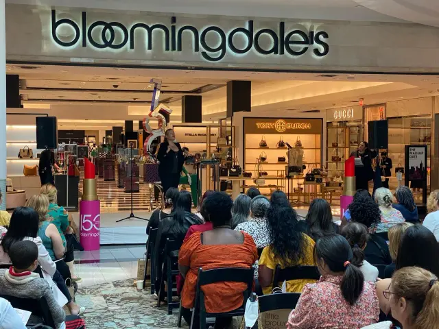 Bloomingdale's store with an event featuring a speaker on stage in front of an audience. The display includes large decorative lipstick and signage for 'The Makeup Date.' Designer brands like Gucci and Tory Burch are visible in the background.