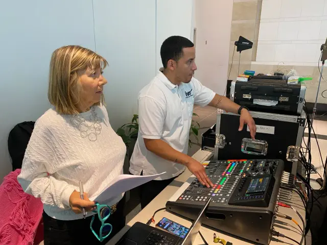 Two people standing at a sound mixing console. One person is holding a paper and glasses, while the other adjusts controls.