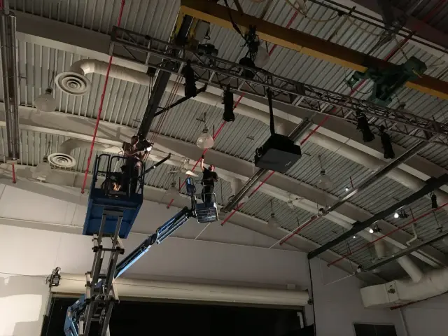 Two workers on a blue scissor lift installing or adjusting lighting equipment on a high ceiling with industrial ductwork.