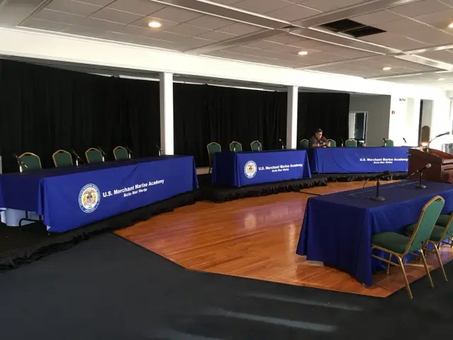 Conference room setup with long tables covered in blue tablecloths labeled "U.S. Merchant Marine Academy" and several green chairs behind them. Microphones are positioned on the tables, and a podium is nearby.