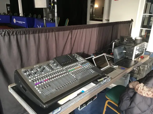 Audio mixing console and laptops on a table backstage at an event, with a viewing panel for adjusting sound levels and other audio equipment.
