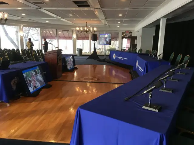 Conference room with long tables covered in blue tablecloths, arranged in a U-shape with microphones on them. Two large screens display images at the front of the room. Chairs are neatly arranged, and a podium is placed in front of the screens. "U.S.