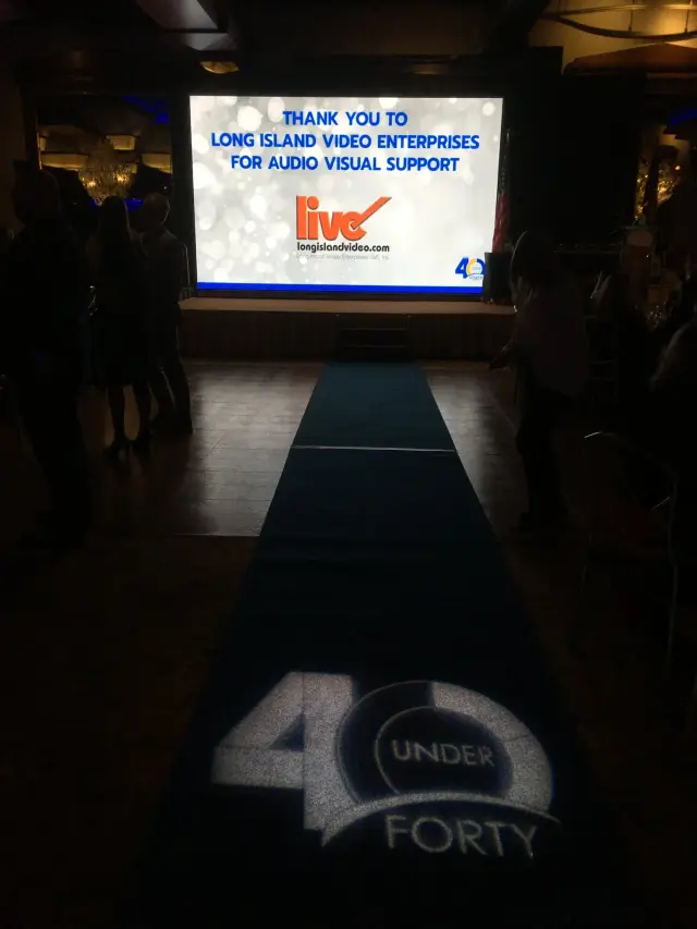 Event stage with a large screen displaying a thank you message to Long Island Video Enterprises for audio visual support at a '40 Under Forty' event, with a projected '40 Under Forty' logo on the floor.