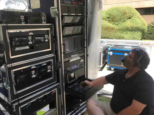 A person wearing a mask and headset operating audio and video equipment in a rack system outdoors.
