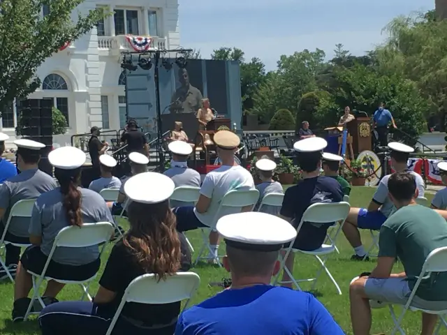 People sitting in folding chairs wearing white uniforms and hats, attending an outdoor ceremony with speakers at a podium.