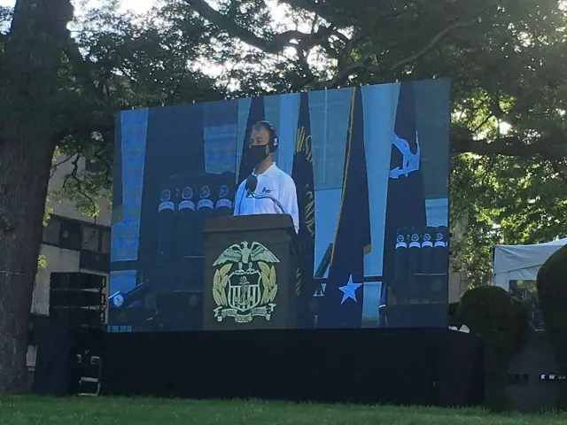 Large outdoor screen displaying a speaker at a podium with a Navy emblem, flags, and trees in the background.