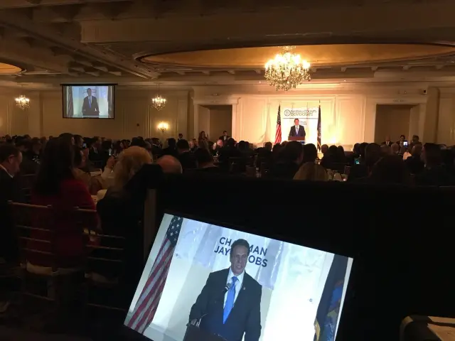 Indoor political event with a speaker on stage and audience seated at tables.