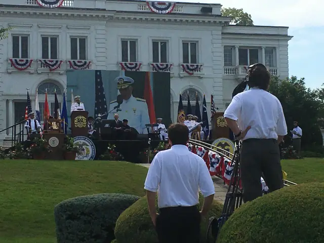 Military ceremony at an outdoor venue with officers in uniform, a podium, flags, and a large outdoor screen displaying a speaker. A cameraman is recording the event.