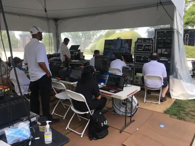 People working in a media production tent with computers, monitors, and technical equipment.