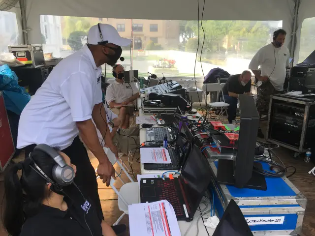 A group of people wearing face masks and headsets working at a tech setup in a tent, with laptops, monitors, and audio equipment on tables.