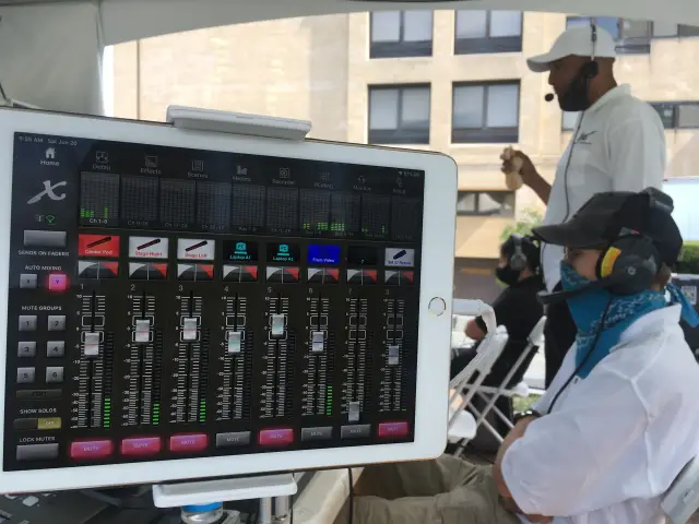 A close-up of a digital audio mixing console on a tablet at an outdoor event with two people wearing communication headsets in the background.
