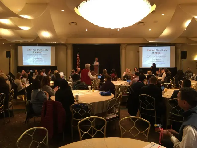 Indoor conference with people sitting at round tables facing a stage with two large screens displaying a presentation titled 'What Are They Really Thinking?'.