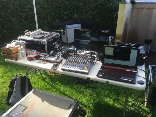 Outdoor video production setup on a table with laptop, video mixer, monitors, cables, and various equipment.