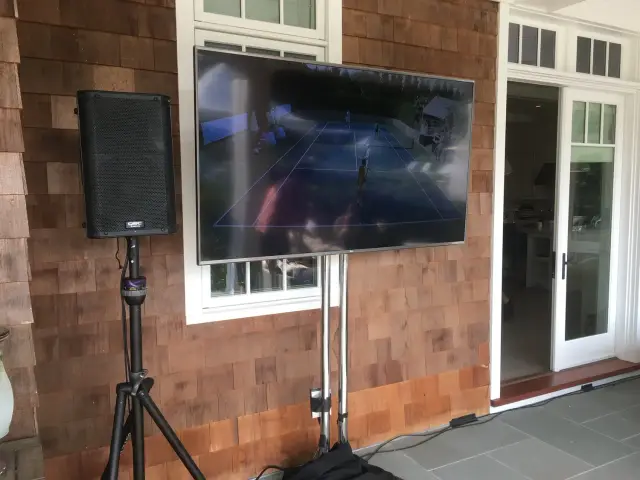 Outdoor setup with a flat-screen TV displaying a tennis match, mounted on a wooden wall next to a professional speaker on a tripod stand.