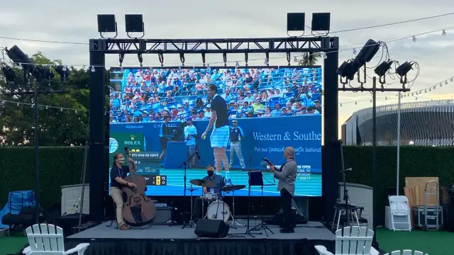 Outdoor event with live band playing in front of a large screen showing a tennis match.