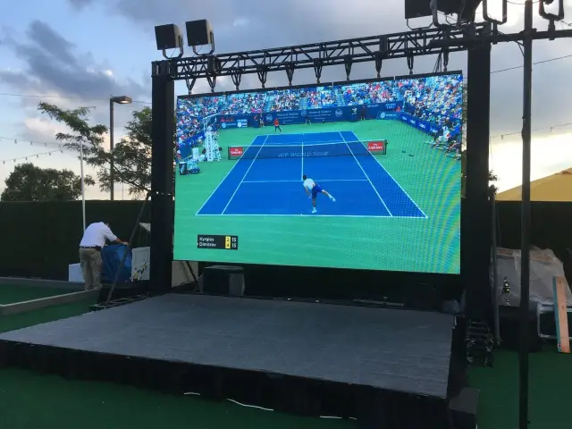 Large outdoor screen showing a live tennis match, featuring a player serving on a blue court. The surroundings include trees, a man in a white shirt adjusting equipment, and a stage with artificial turf.