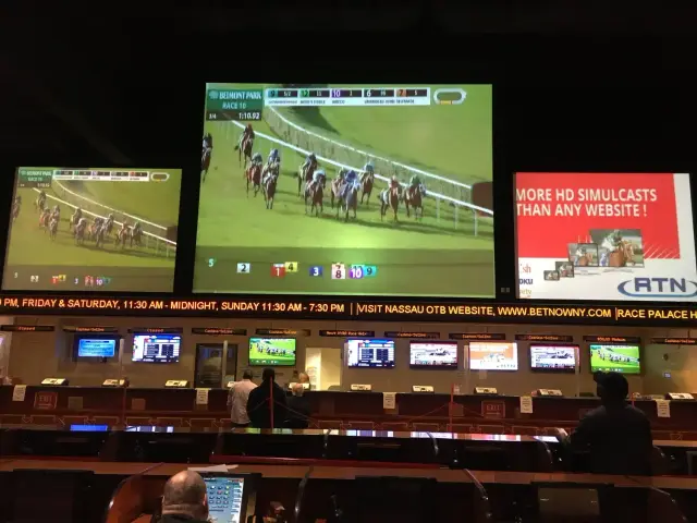 Interior of a betting venue with large screens showing a live horse race from Belmont Park. Digital displays and betting terminals are visible below, with people watching the event.