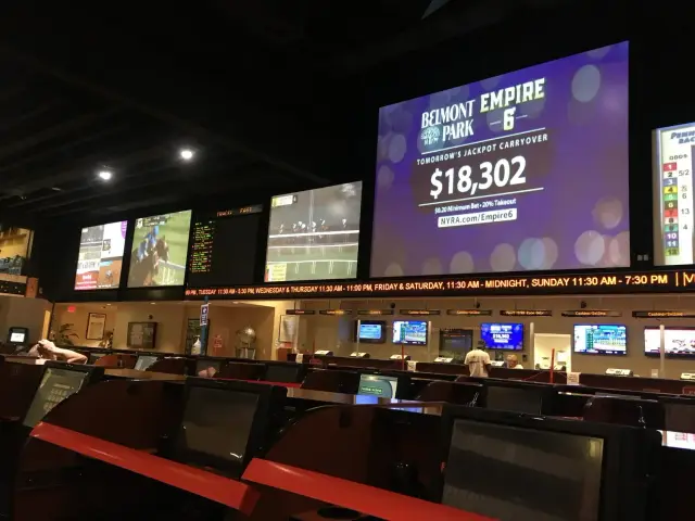 Interior of a sports betting facility with large screens displaying horse racing and betting information. People are seated at betting terminals, and a digital ticker shows operating hours. The main screen advertises a jackpot at Belmont Park.