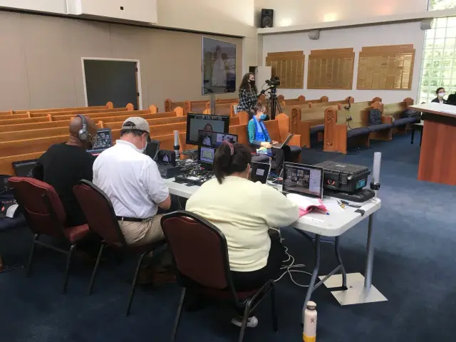 People in a church with audiovisual equipment, managing a live stream or recording. Empty pews are visible in the background.