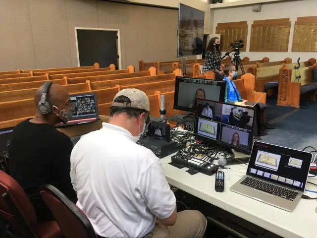 Two men sit at a table with recording and broadcasting equipment in a church setting, where a woman operates a camera. Monitors display virtual meeting with participants.