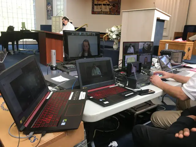 Broadcast setup at an event with multiple laptops and screens showing a speaker delivering a speech, monitored by a technician.