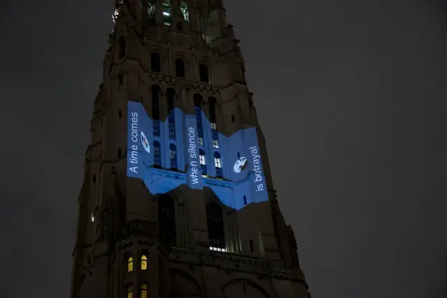 A tall building with text projected on its facade at night reads, "A time comes when silence is betrayal." There are windows and architectural details visible, illuminated by the projection.