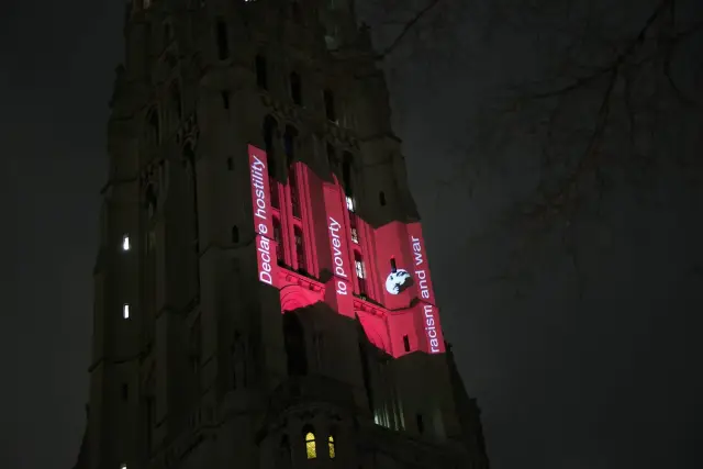 Church tower illuminated with red projections and anti-poverty, racism, and war messages, including a silhouette of a man's face.