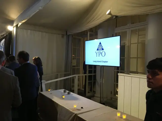 People gathered at an event with a sign displaying 'YPO Long Island Chapter' on a screen, next to tables with candles.