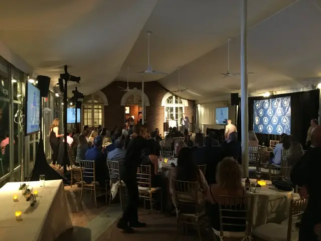 Indoor event with audience seated in front of a stage, under a large tent, featuring a speaker and a backdrop with logos. Tables with candles are visible, and a cameraman is present.