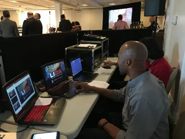 People working on laptops and equipment at a tech setup in a busy conference room, with a screen in the background.